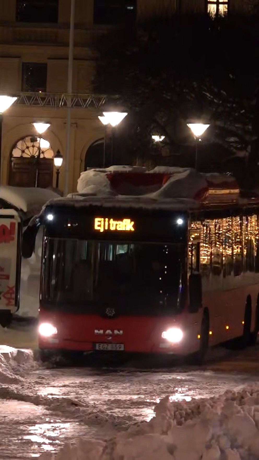 Två stadsbussar står parkerade vid en hållplats i snöigt vinterlandskap, med belysning från gatlampor och omgivande byggnader i bakgrunden. En person går förbi hållplatsen, och en buss har texten ”Ej trafik” synlig, medan den andra har linjenumret ”14” angivet.