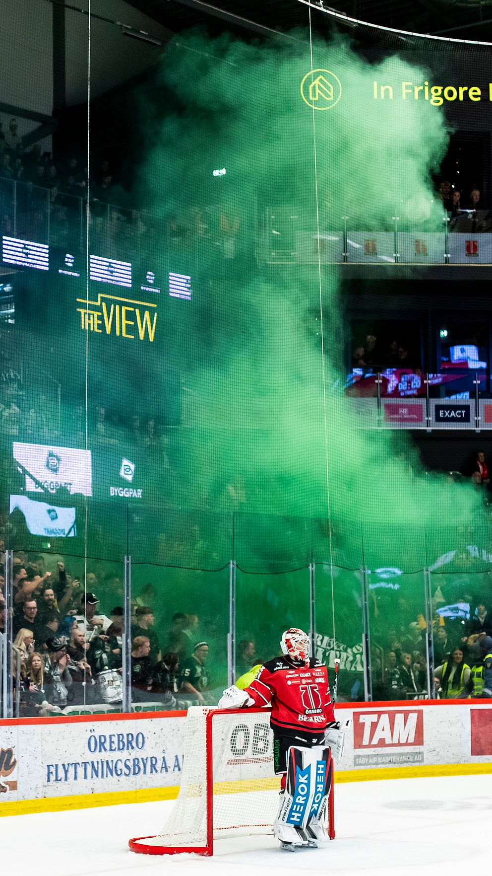 En hockeymålvakt i röd utrustning står framför målet medan gröna rökpelare sprider sig bakom honom, vilket skapar en dramatisk stämning. Publiken är entusiastisk och synlig i bakgrunden, omgiven av reklamskyltar och ljus.