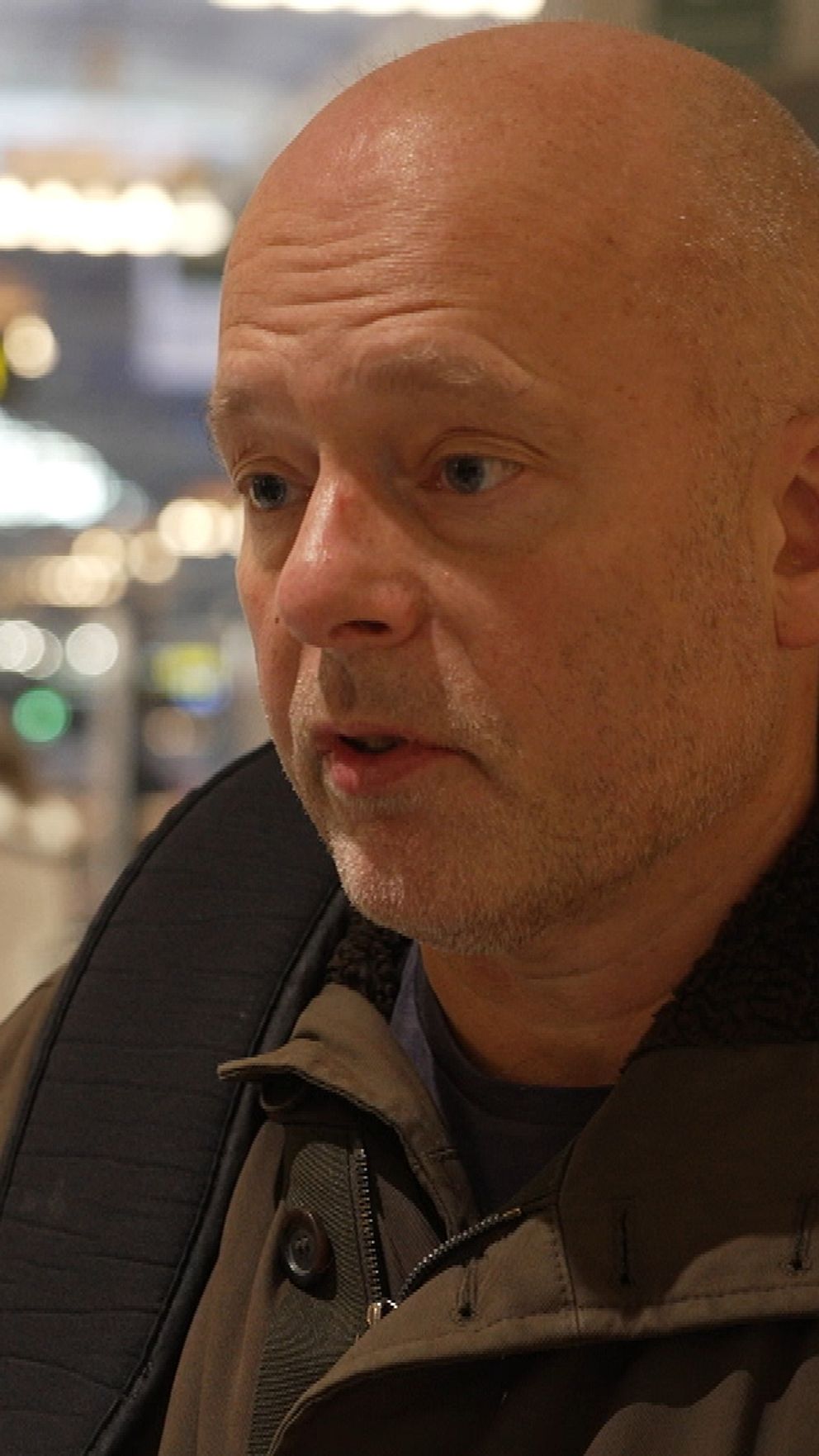 En man i vinterkläder står på en livlig tågstation med människor i bakgrunden som går förbi och informationstavlor som visar tågtider.