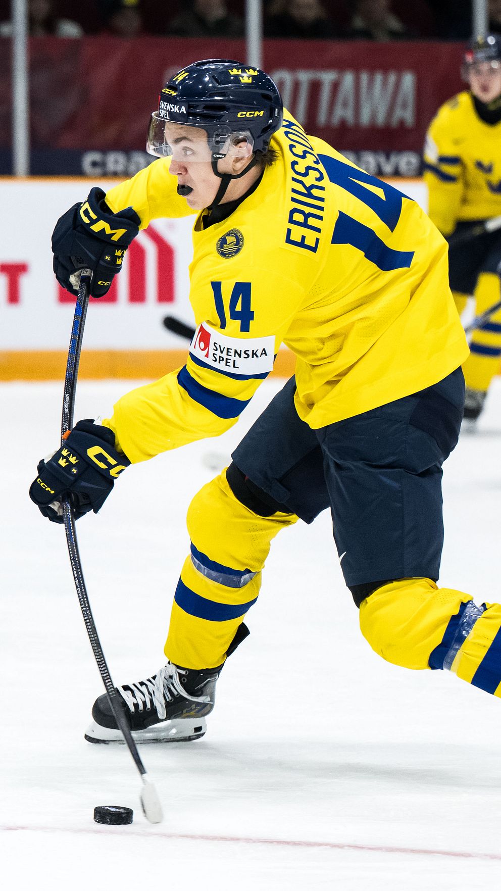 Svensk hockeyspelare med tröja nummer 14, Eriksson, åker framåt med pucken under en match. I bakgrunden syns en motståndarlagsspelare på väg in i spel. Arenan har tydligt skyltar och en entusiastisk publik.