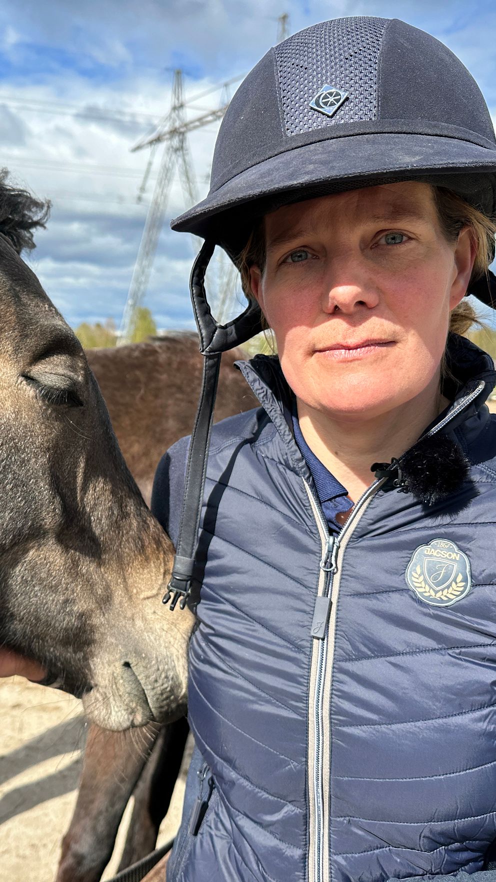 En person med ridhjälm står nära en häst som nosar mot personens axel utomhus.