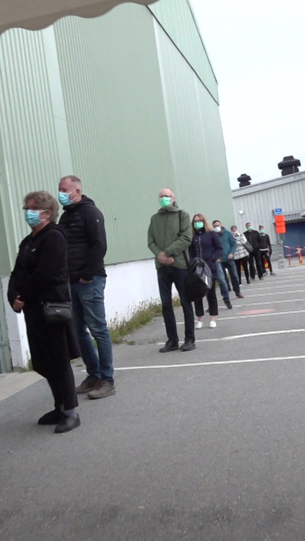 Människor står på rad utomhus, många med ansiktsmasker, väntande på att komma in i ett större grått byggnadskomplex. Avspärrningar skapar en tydlig köled.