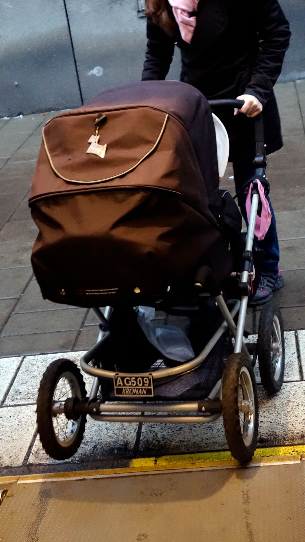 En kvinna kliver ut ur en buss med en barnvagn. Barnvagnen är av mörk färg och är placerad på trappan som leder ner från bussen. Kvinna bär en svart jacka och ser ut att hjälpa barnet i vagnen. Området omkring är asfalt, vilket tyder på en urban miljö.