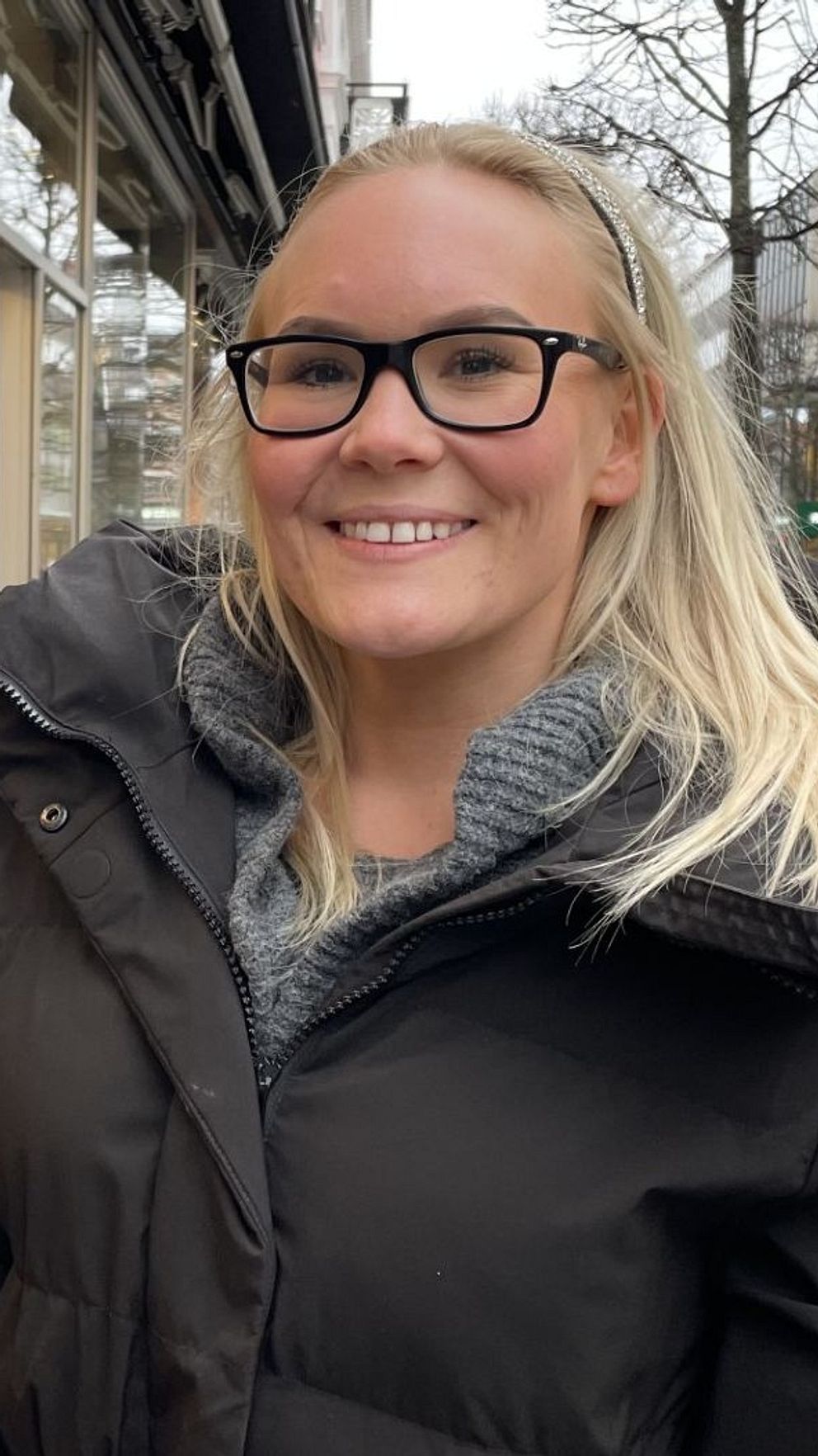 En kvinna i en svart vinterjacka står på en trottoar i en stadsmiljö med glasfasader och butiker på ena sidan. I bakgrunden syns människor som går längs gatan, samt träd och gatubelysning.