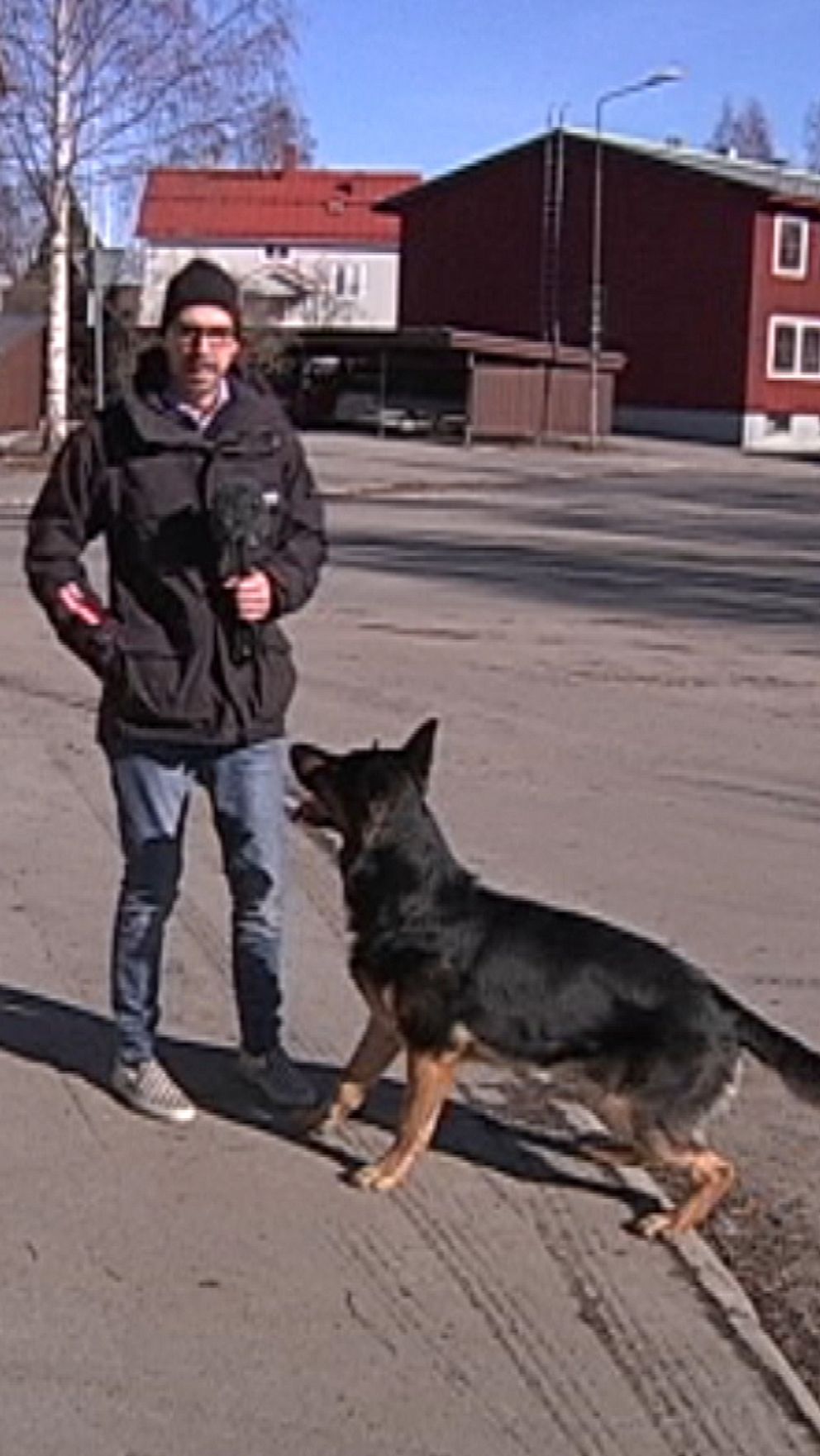 Man promenerar med hund på solig stadsöppen plats, bostadshus i bakgrunden.