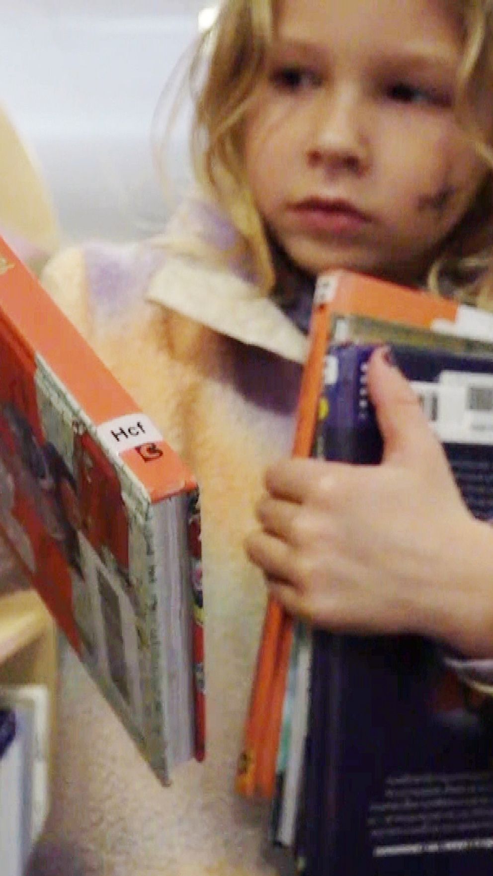 Ett barn plockar böcker från en bokhylla, med flera böcker i den ena armen och en annan bok i den andra handen. Barnet har på sig en mjuk, flerfärgad jacka och står inuti en biblioteksmiljö med synliga bokhyllor i bakgrunden.