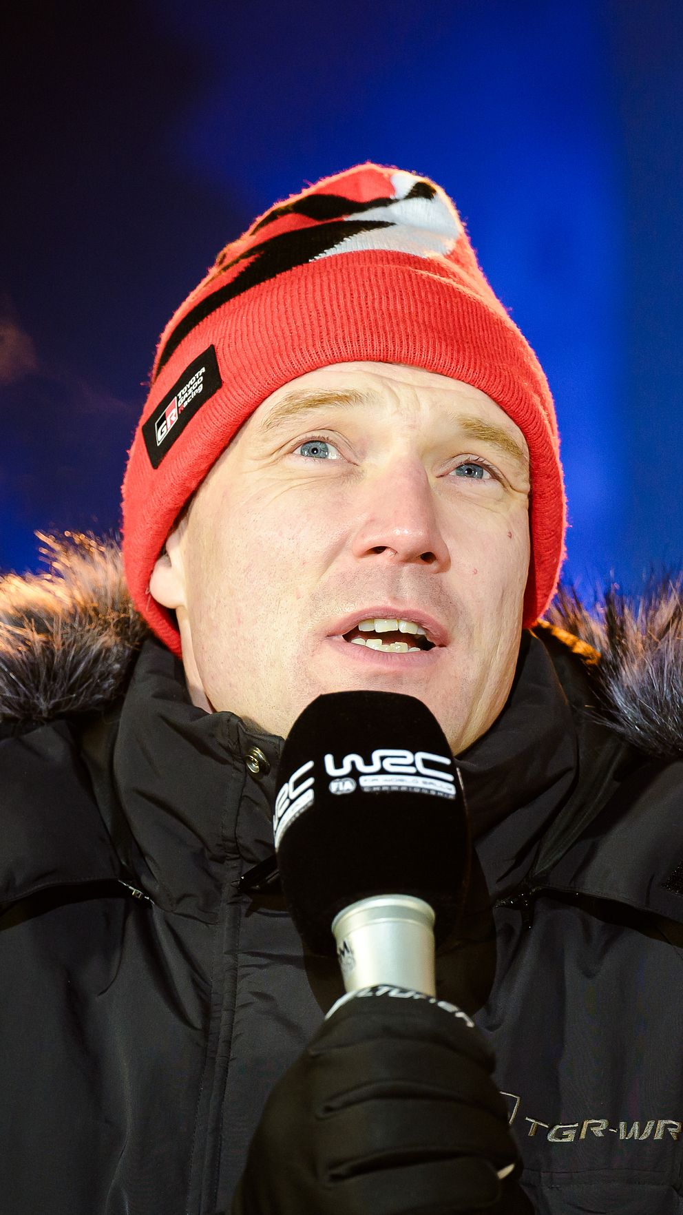Jari-Matti Latvala i vinterkläder håller en mikrofon med ånga som stiger upp i den kalla luften, mot en suddig bakgrund med ljusa färger och logotyper.