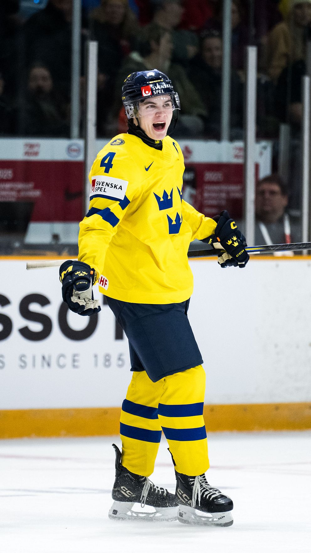 Två svenska ishockeyspelare firar ett mål på isen, iförda gula och blå tröjor med nummer 2 och 4. I bakgrunden syns publiken och en stor klocka, vilket ger en känsla av en livlig sportsammanhang.