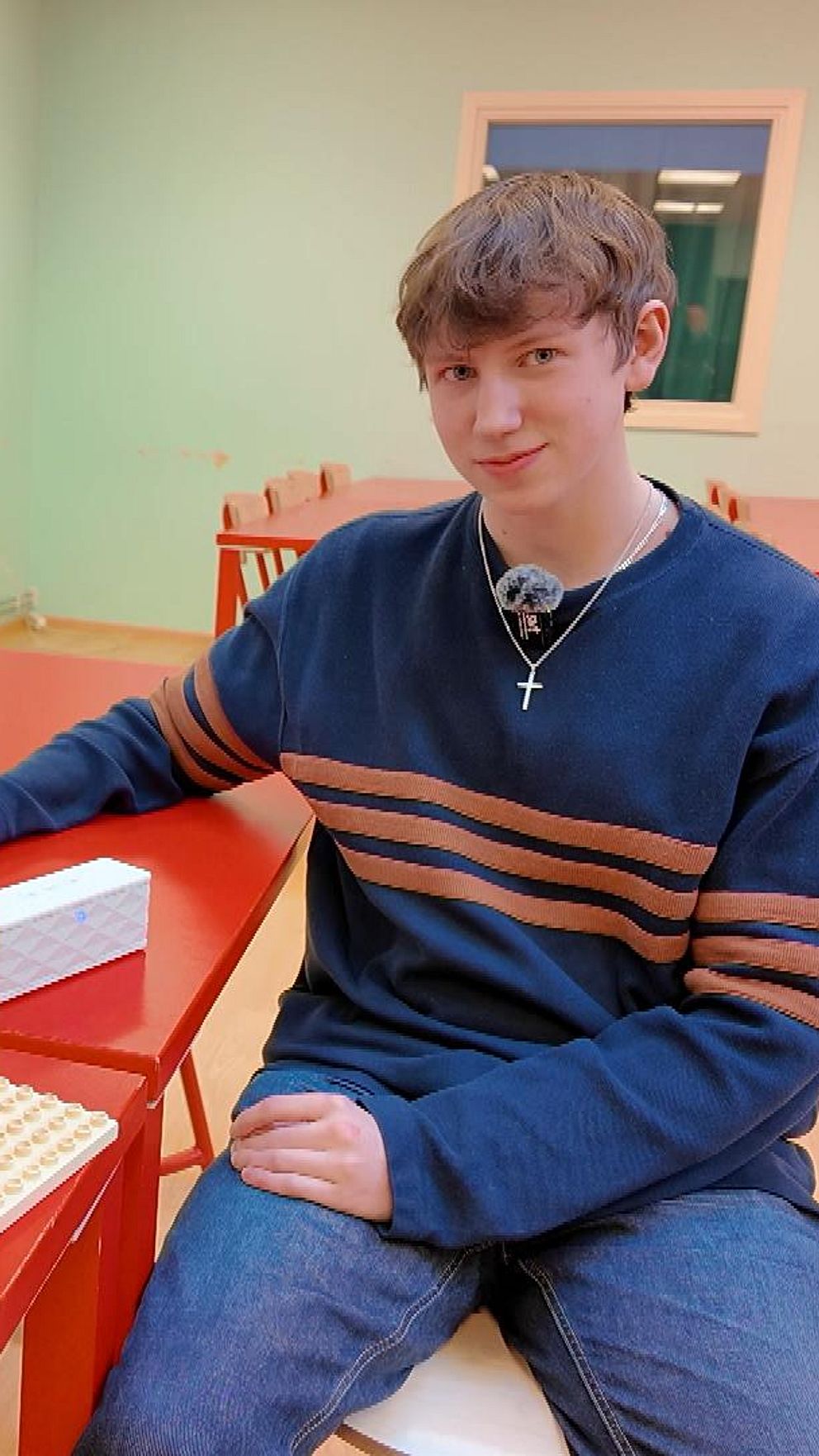 En person sitter vid ett rött bord i ett klassrum och interagerar med en elektronisk enhet som är monterad på ett stativ av LEGO-bitar.