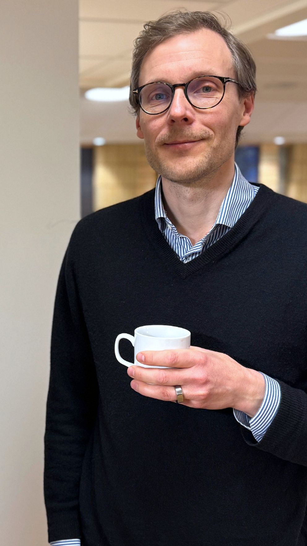 Person står bredvid en kaffemaskin, håller en liten kaffekopp. Bakgrund med bord och stolar.