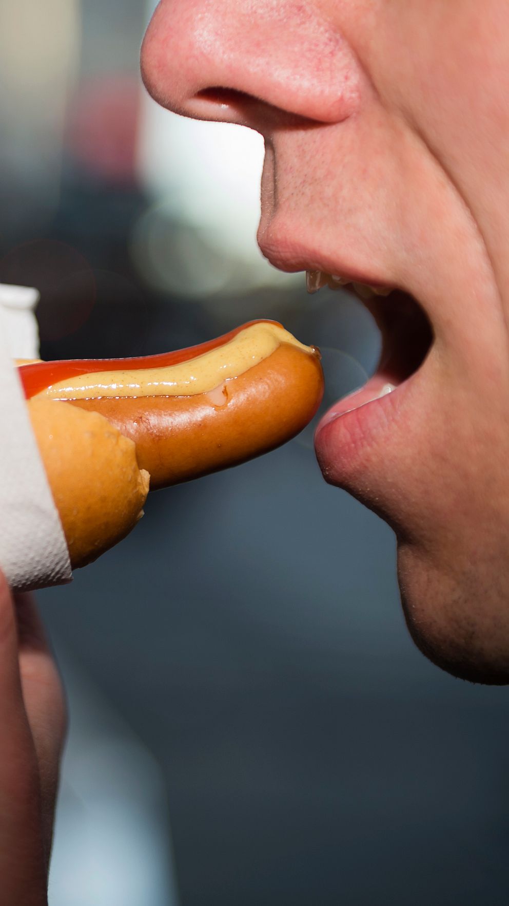 En hand håller en varmkorv insvept i en pappersservett, med ketchup synlig på toppen. Bakgrunden är suddig och visar en stadsmiljö i skymning.