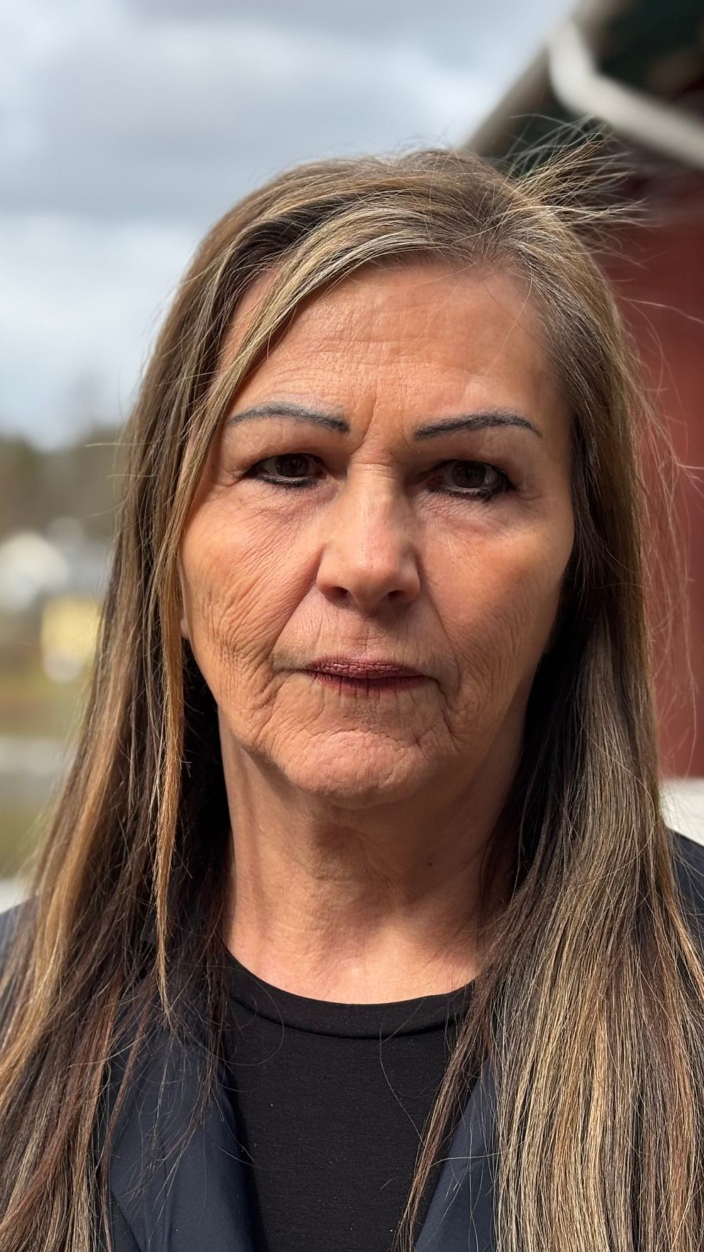 Kvinna med långt hår står utomhus vid ett hus, bostadsområde syns i bakgrunden.