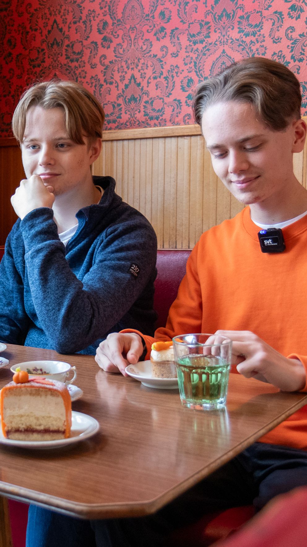Tre personer sitter vid ett bord på ett café. De har bakverk och drycker framför sig. En person håller en telefon och tar ett foto, medan de andra verkar prata. Rummet har en varm och inbjudande atmosfär med röd tapet och träpaneler.