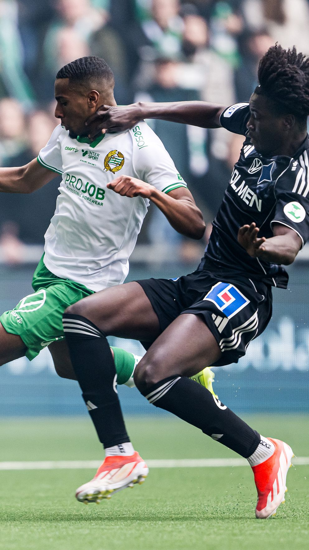 Två fotbollsspelare kämpar om bollen under en match med publik i bakgrunden.