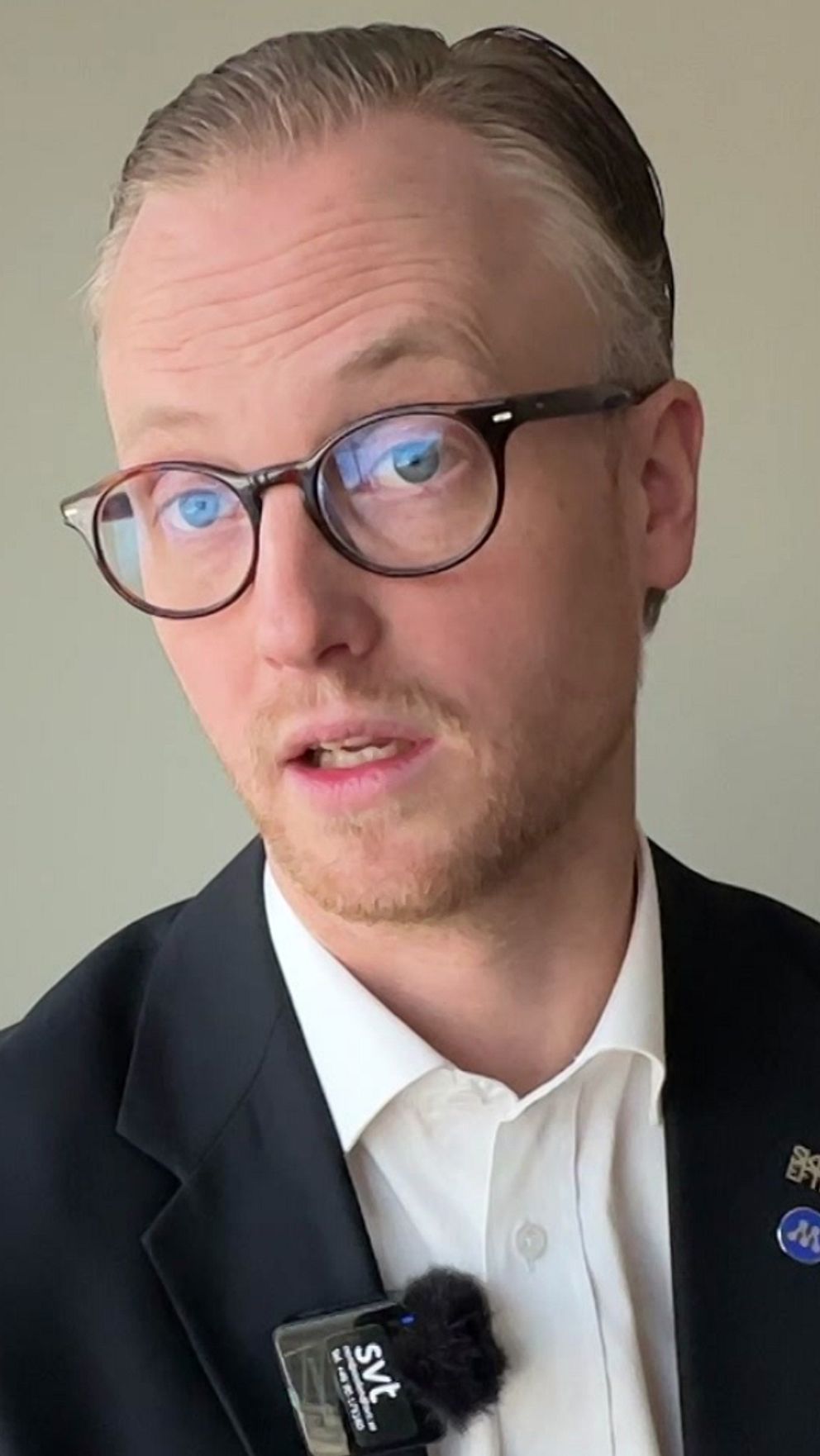 Person i kostym står vid ett fönster. Mikrofon på kavajen.