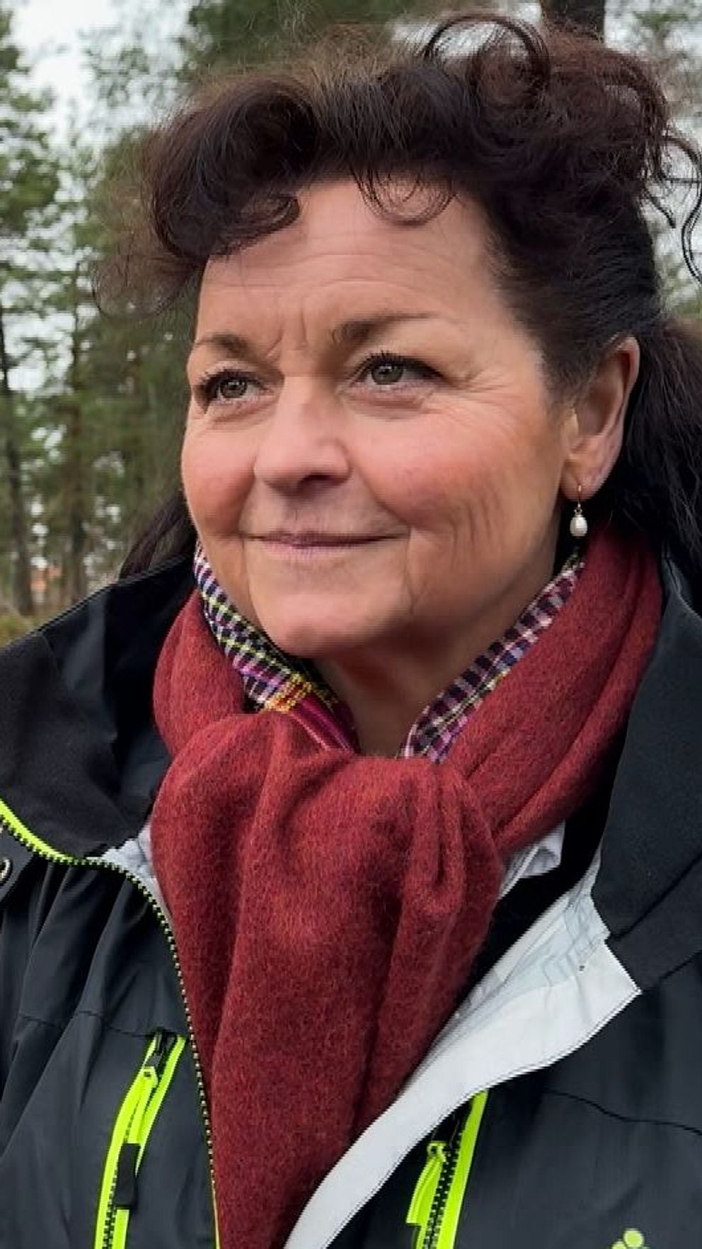 En person står i en skogsmiljö, klädd i en mörk jacka med ljusa detaljer och en röd halsduk. Skogen består av höga tallar och marken är täckt av mossa och låg vegetation.