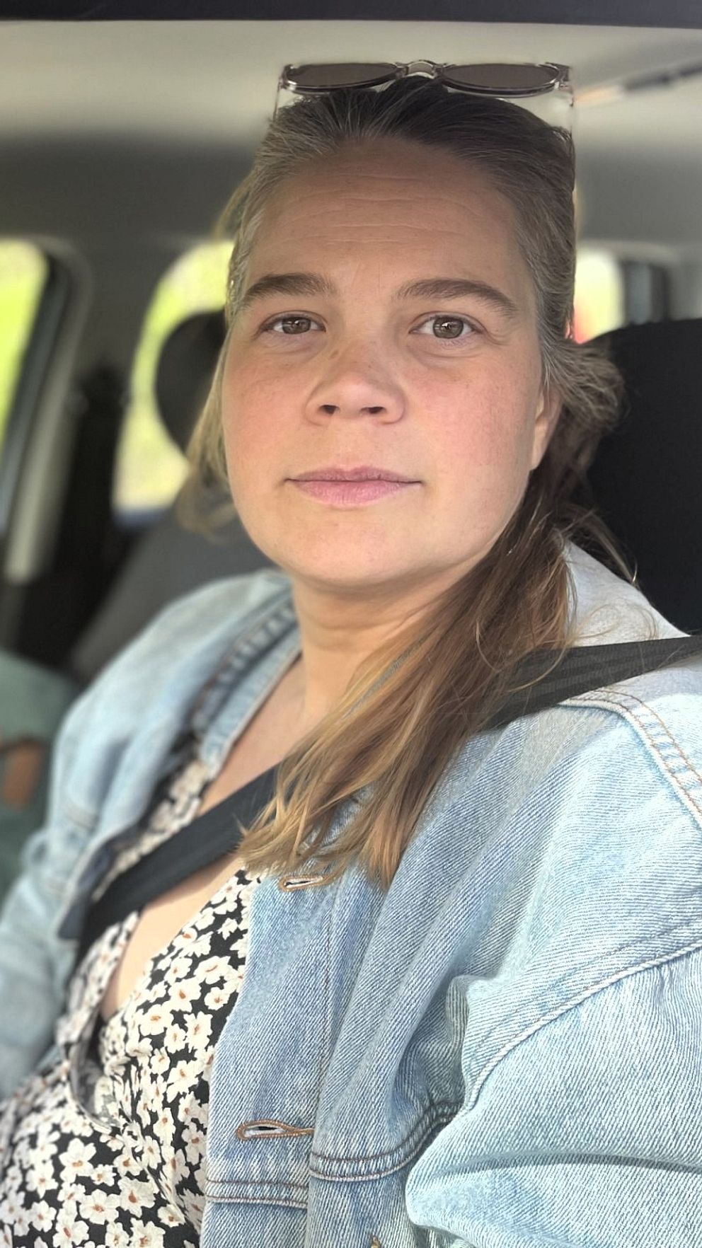 Person sitter i bil med säkerhetsbälte, omgiven av grönskande bakgrund utanför fönstret.