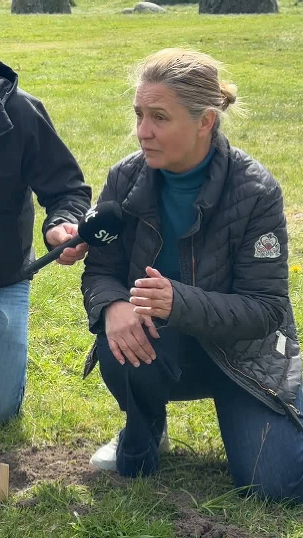 Två personer undersöker marken intill en stor sten på en gräsyta utomhus.