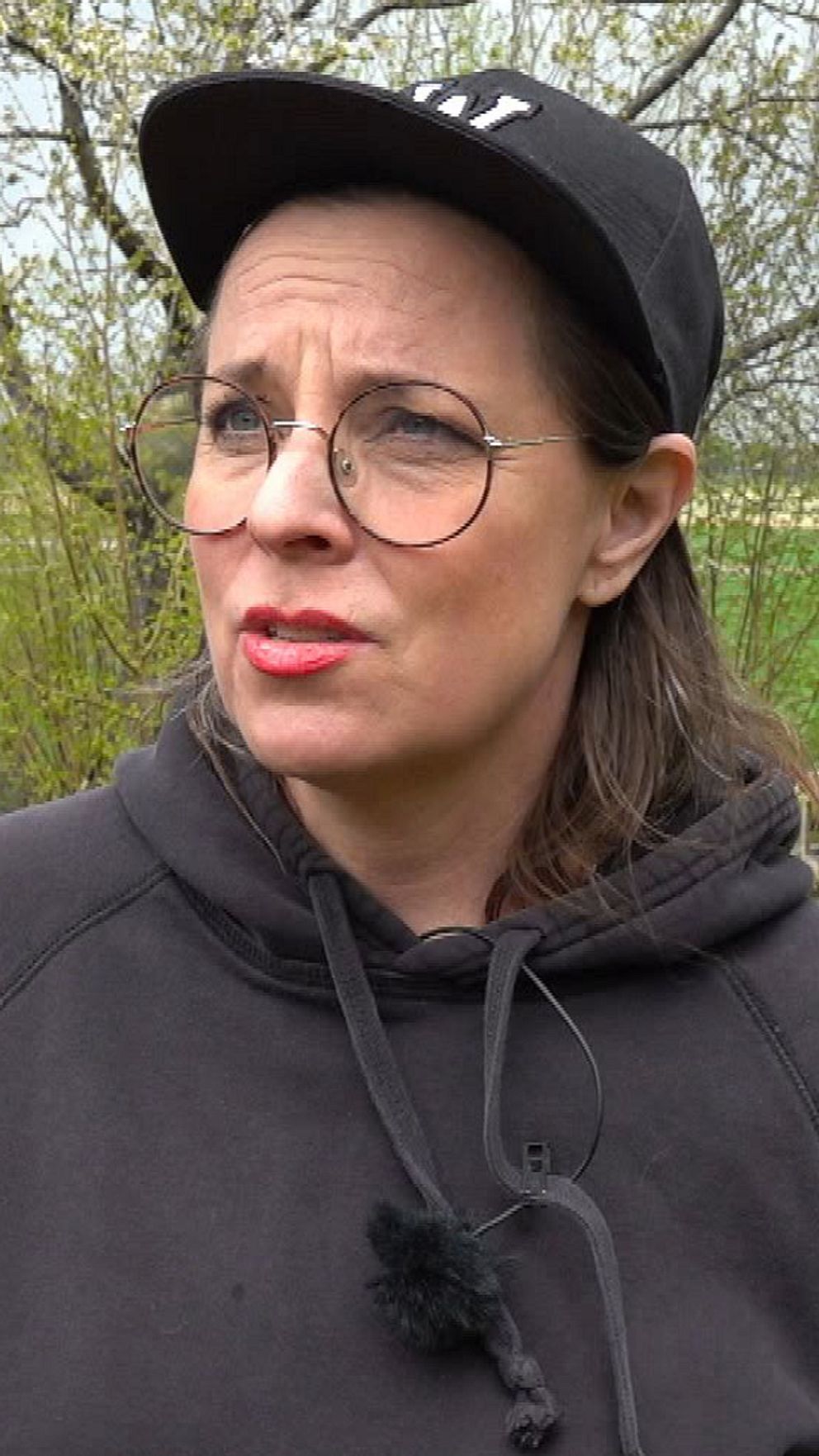 Person med keps utanför ett rött hus med trädgårdsslang och grönska i bakgrunden.