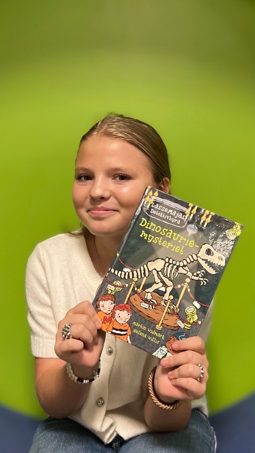 En person håller upp en bok med titeln ”Dinosaurier-mysterier” i framkant, med en färgglad bakgrund i grön nyans. Boken innehåller en illustration av ett dinosaurieskelett och två barn på omslaget.