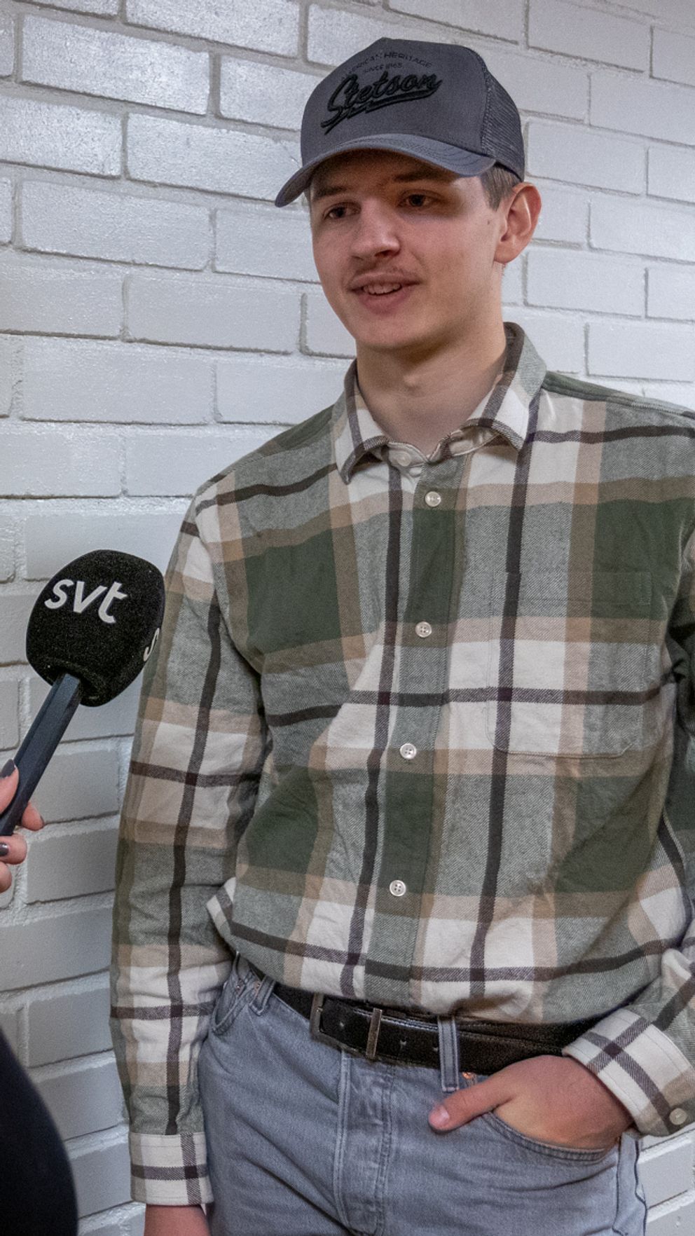 En person i en rutig skjorta intervjuas av en journalist som håller en mikrofon med SVT-logotyp. Bakgrunden består av vit tegelvägg. Journalisten har långt, rödaktigt hår och vänder sig mot den intervjuade personen.