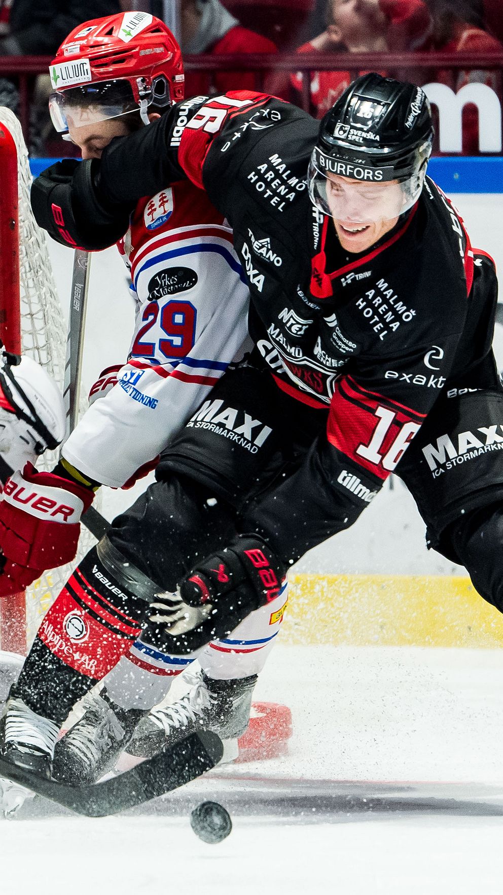 Ishockeyspelare från två olika lag kämpar om pucken nära målet. Målvakten i svarta kläder försöker avvärja ett skott, medan en medspelare skyndar till undsättning. En spelare från det andra laget i vit tröja är också involverad i situationen.