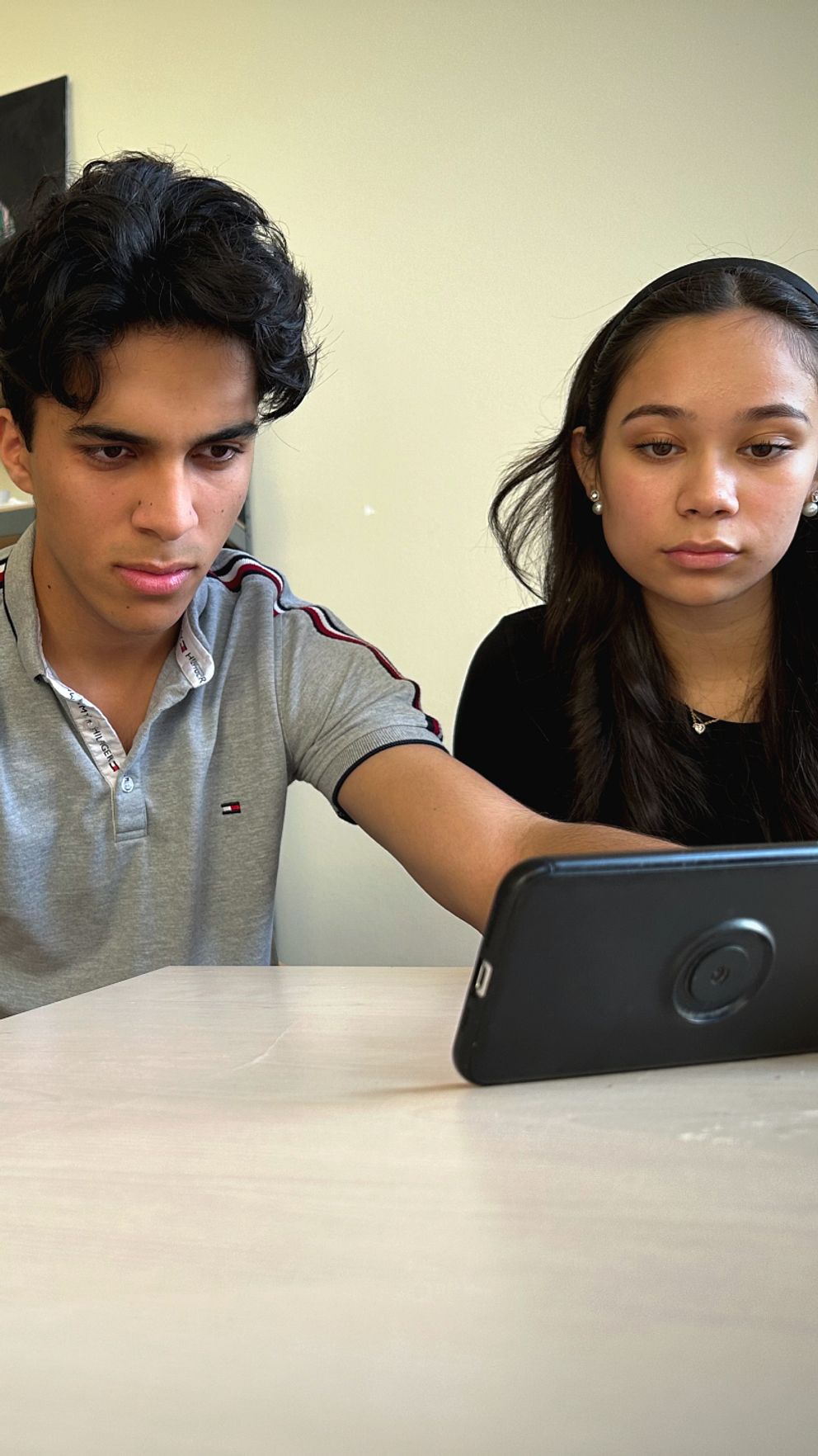 Nadine, Hussameddin och Jenny sitter vid ett bord och tittar på en smartphone. De verkar vara fokuserade på skärmen. Rummet har en neutral bakgrund med en hylla synlig i bakgrunden.