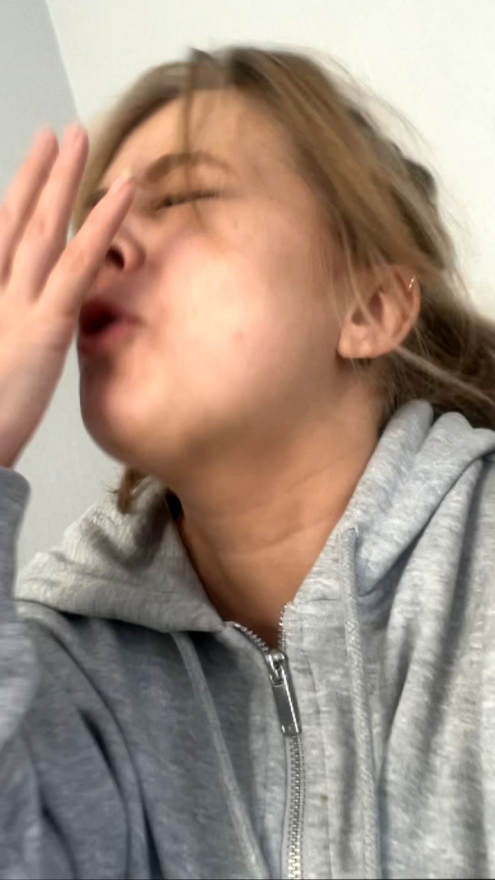 Lovisa med grå huvtröja sitter mot en ljus vägg och håller upp en hand nära ansiktet.