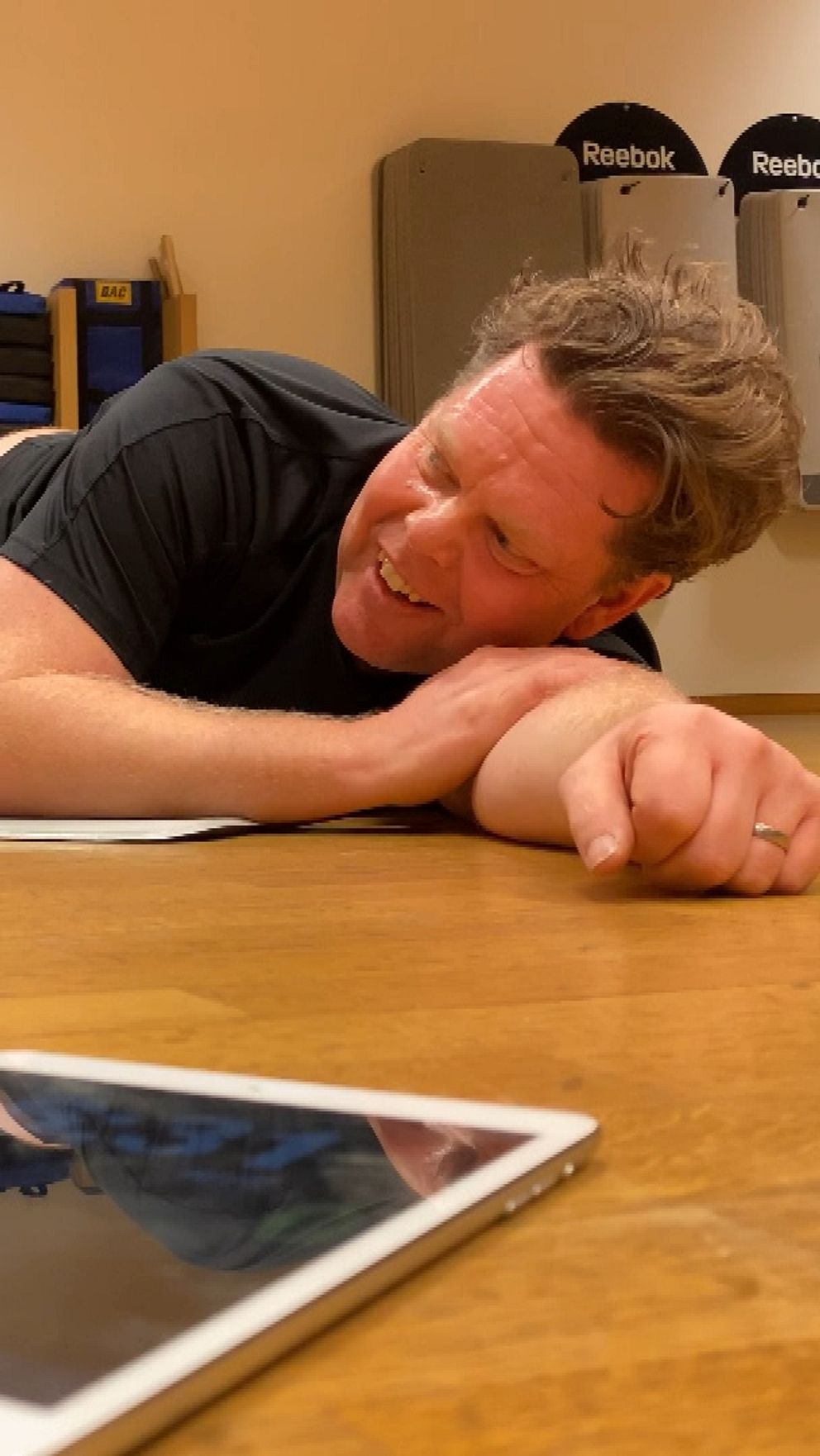 En person ligger ner på en träningsmatta i en gymmiljö, med träningsredskap som vikter synliga i bakgrunden. I förgrunden ligger en surfplatta på golvet, och en bebis sitter nära en hantel.