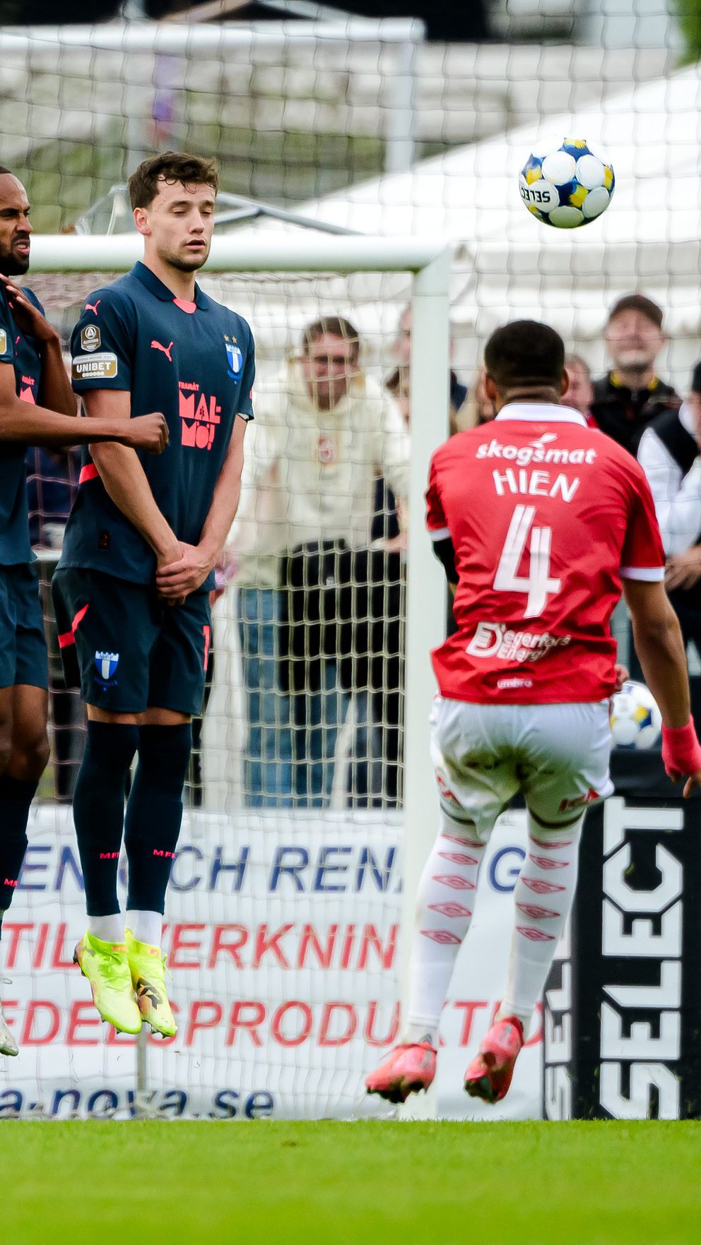Spelare skjuter frispark mot mål, motståndare hoppar i mur och målvakt följer bollen.