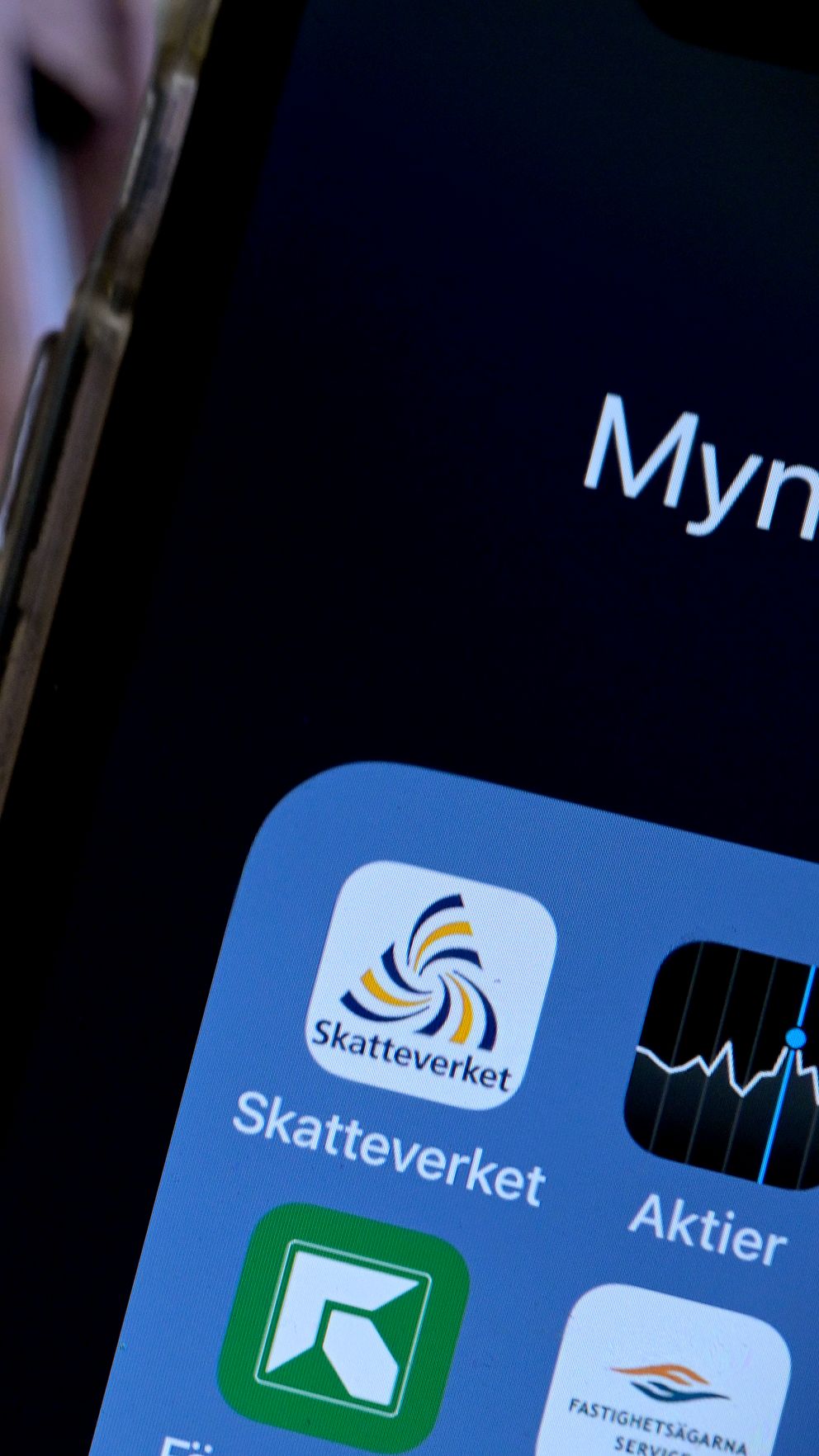 En person håller en smartphone som visar appar med Skatteverkets logotyp i förgrunden. I bakgrunden syns Skatteverkets logotyp och namn på en byggnad.