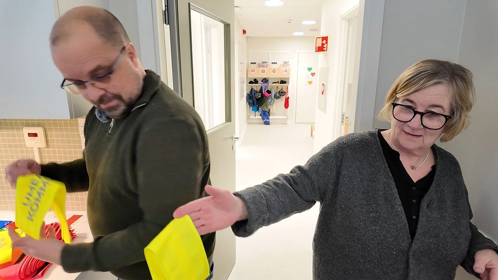 Två personer står i en ljus korridor med dörröppningar i bakgrunden. Den manliga personen till vänster håller en gul armbindel, medan den kvinnliga personen till höger sträcker fram en annan gul armbindel. Korridoren är inredd med vita väggar och det syns klädhängare med flerfärgade kläder i bakgrunden.