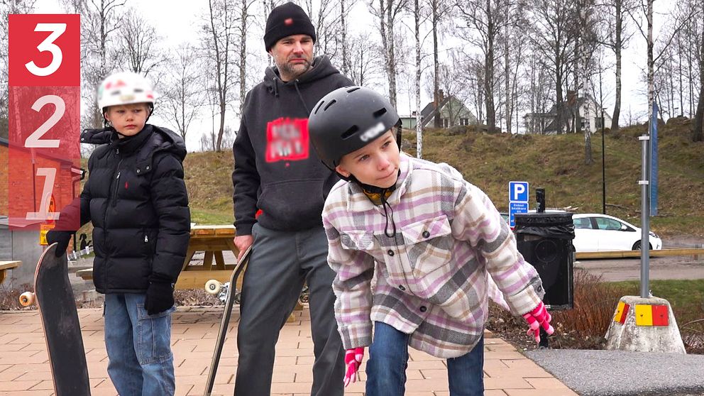 Två barn med hjälm och skateboard i skatepark tillsammans med en vuxen person.