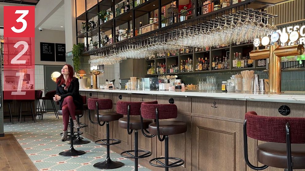 En stilren bar med en lång bänkdisk, inredd med trä och metall. Hyllor med alkoholflaskor syns bakom disken, där glas hänger i rader. Emma Engdahl, kärleksexpert, sitter på en barstol klädd i svarta kläder, vänd mot kameran. Golvet är dekorerat med ett mönstrat kakel och det finns växter i lokalen som bidrar till den moderna atmosfären.