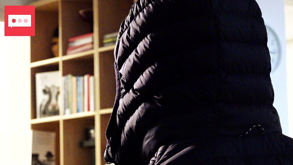 En person iförd en svart dunjacka med huva står vänd bort från kameran i ett rum. I bakgrunden syns en bokhylla med olika böcker och prydnadsföremål.