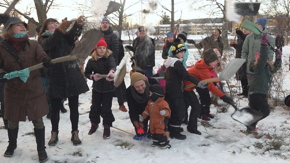En grupp människor, inklusive barn, deltar entusiastiskt i en snöbollskastning eller lek i ett snötäckt parkområde. Några i gruppen använder spadar för att hantera snön. Stämningen verkar glad och livlig med en bakgrund av träd och en byggnad i fjärran.
