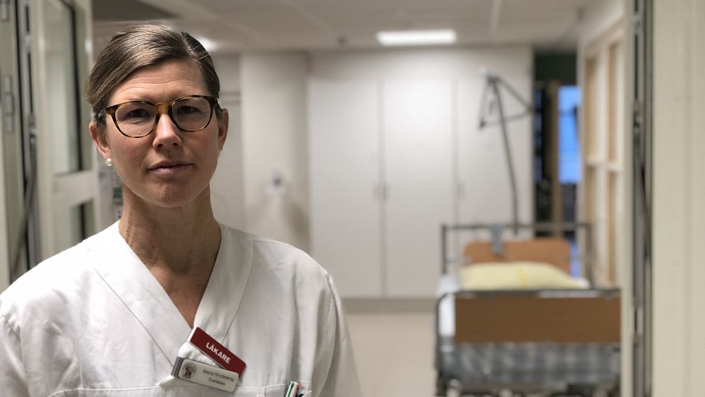 En läkare i vit uniform står i en sjukhuskorridor. Bakgrunden visar en tom sjukhussäng med sängkläder, vilket indikerar en läkarmiljö.