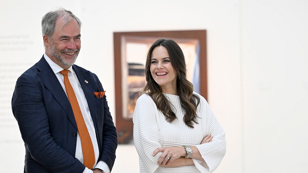 Två personer står tillsammans i en konstutställning, med en målning i bakgrunden. Den ena personen bär en mörk kostym med en orange slips, medan den andra är klädd i en vit, randig tröja och har armarna i kors. Atmosfären ser ut att vara avslappnad och konversationsinriktad.