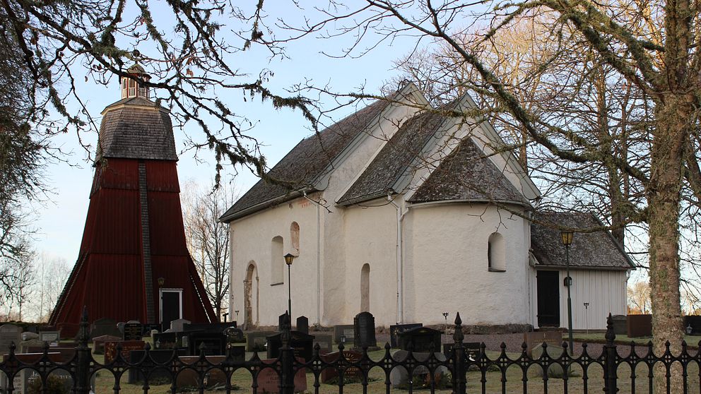 Vit stenkyrka med röd klockstapel i förgrunden, omgiven av träd och gravstenar vid en kyrkogård.