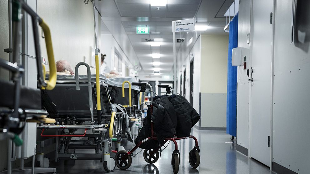 Sjukhuskorridor med sjuksäng där man ser en mans bakhuvud och en rullator vid sidan om sängen