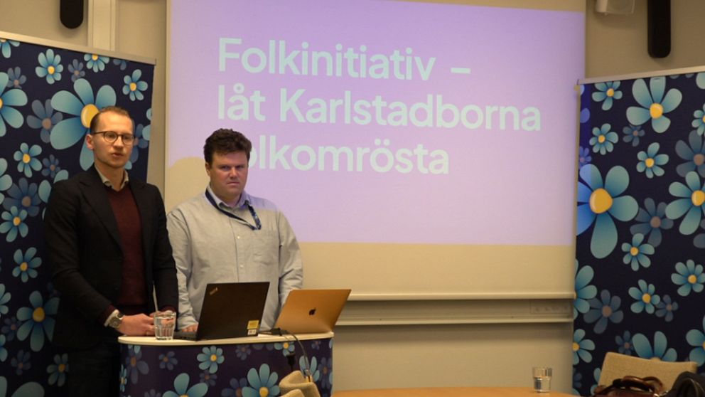 Två personer står vid ett podium med SD-blommor med en presentation om ”Folkinitiativ – låt Karlstadborna folkomrösta” i bakgrunden.