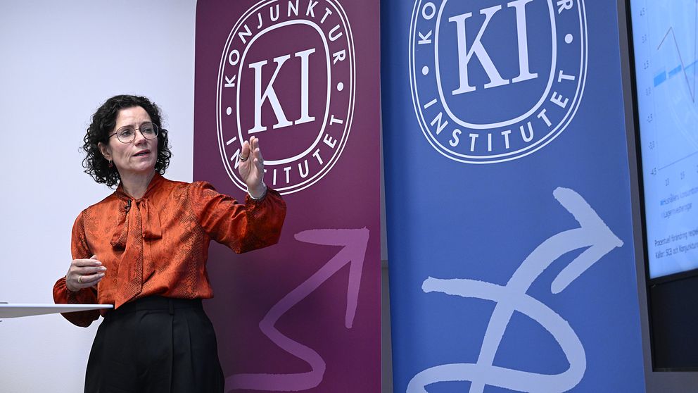 En person står och presenterar framför två banner med logotyper för Konjunkturinstitutet, en i röd och en i blå färg.