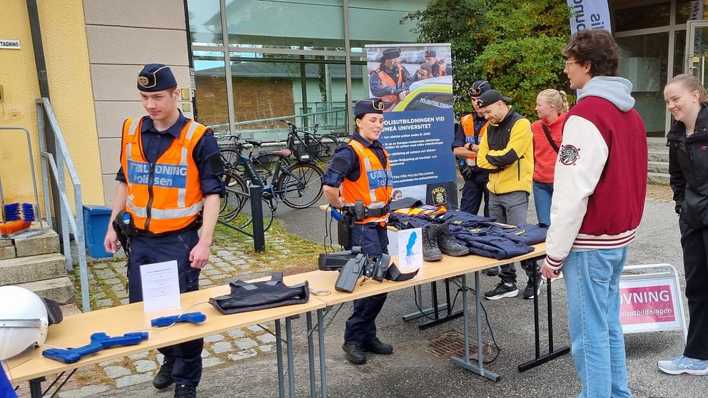 Två polisstudenter står bakom ett bord och en man med röd jacka står framför