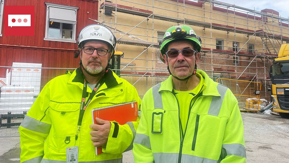 Två personer i reflexvästar står på en byggarbetsplats. Den ena har en skrivbok i handen och båda bär hjälmar med belysning. I bakgrunden syns en byggnad under konstruktion med byggställningar. En lastbil står parkerad i närheten.