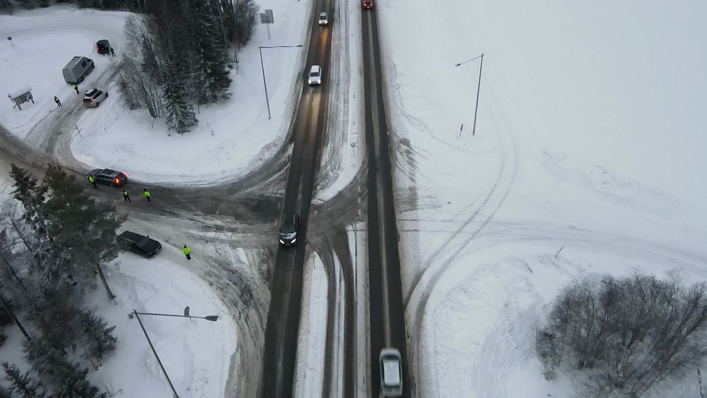 Vy av en trafikerad korsning under vinterförhållanden, där snö täcker vägen och omgivande landskap. Flera fordon rör sig genom korsningen, och två personer i reflexkläder står vid sidan av vägen. Området omges av träd och en sjö syns i bakgrunden.