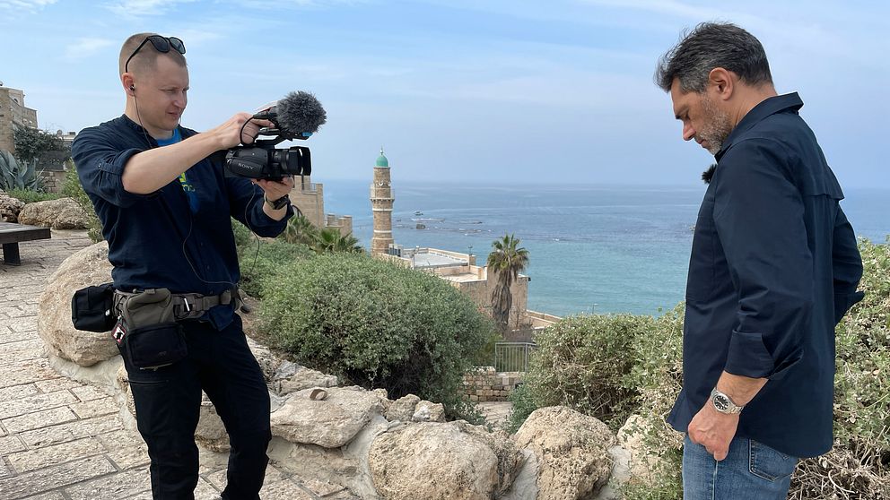 Samir Abu Eid står på en stenlagd promenad med utsikt över havet, medan Johannes Tolf filmar med en kamera. I bakgrunden syns en historisk byggnad och en utsiktstorn, med palmer och havet i horisonten.