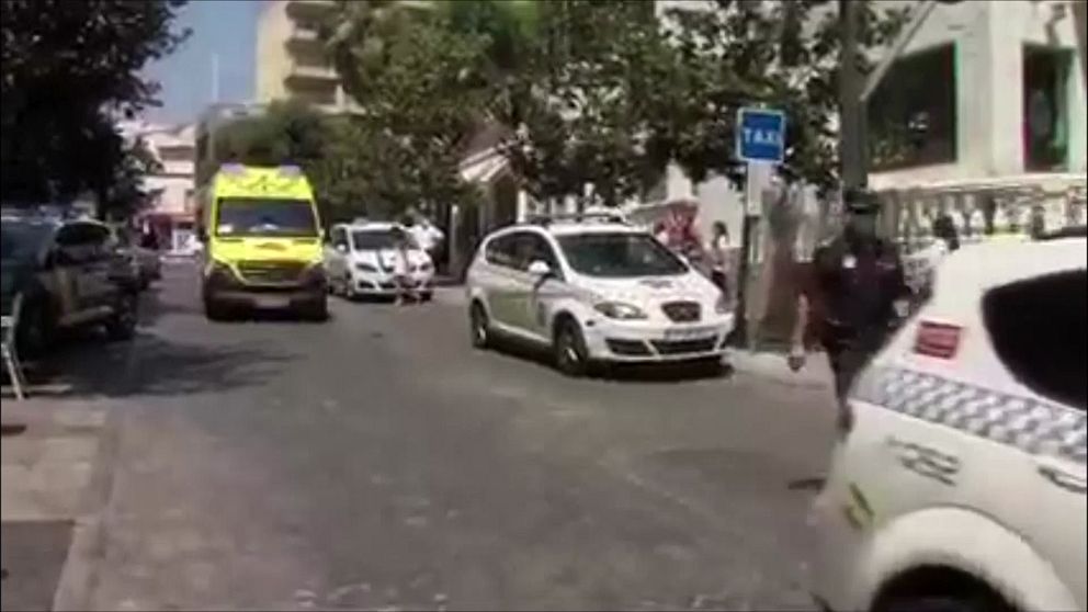 En gata med flera fordon, inklusive en ambulans och polisfordon, som rör sig bland träd och byggnader. I bakgrunden finns en skylt för taxi, och några personer syns på trottoaren.