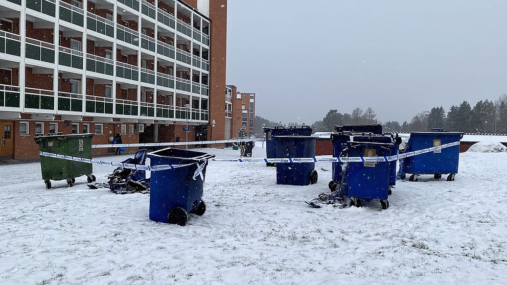 Sju soptunnor i olika färger, inklusive gröna och blå, står på en snötäckt gräsmatta framför en flerfamiljsbyggnad i stadsdelen Lugnvik i Östersund. Området är avspärrat med blå-vita polisband, och snö faller lätt. Bakom byggnaden syns träd i bakgrunden.