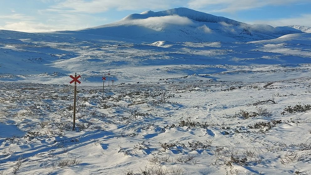 Vy mot Helagsmassivet. Tunt snötäcke. På bilden synsett rött ledkryss som markerar en vinterled i förgrunden och bergen i bakgrunden under en klarblå himmel.