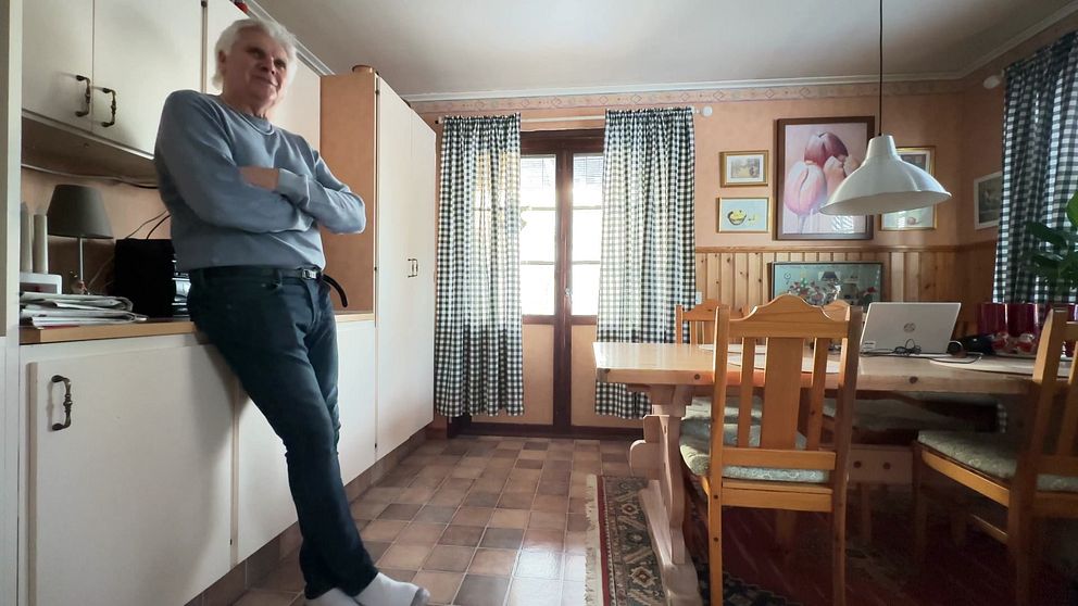 Köksmiljö med ett matbord och stolar i trä, samt ett fönster med rutiga gardiner. I bilden syns en person som lutar sig mot en köksskåp. På väggen hänger några tavlor. Golvet är beklätt med plattor och en matta ligger under matbordet.